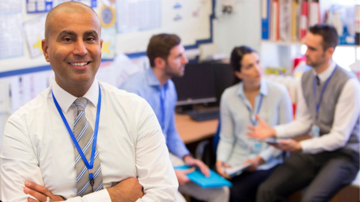 A confident school administrator with crossed arms smiling at the camera, with a background of educators in a collaborative meeting discussing school tech solutions.